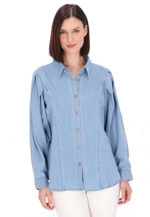 Woman wearing a light blue button-up denim shirt with long sleeves and white pants, standing against a white background.