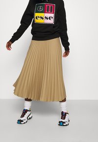 Záhybová béžová sukně v volném designu spárovaná s černou mikinou s barevným grafickým potiskem a bílými, vzorovanými teniskami.