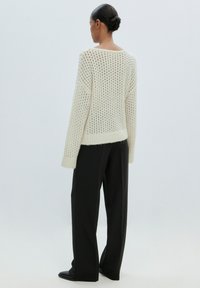 Cremfarbener Häkelstrickpullover mit offener Maschenstruktur, gerippten Bündchen und Saum, kombiniert mit schwarzen, taillierten Hosen und spitzen Schuhen.