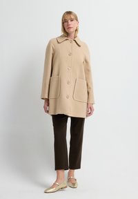 Beige wollen jas met een kraag, vijf grote knopen, twee zakken aan de voorkant en geribbelde details op de manchetten; gecombineerd met een paarse broek en gouden ballerina's.