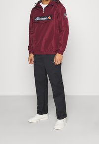 Maroon windbreaker with a quarter zip, large front pocket, and black logo. Paired with black pants and white sneakers. Smooth texture.