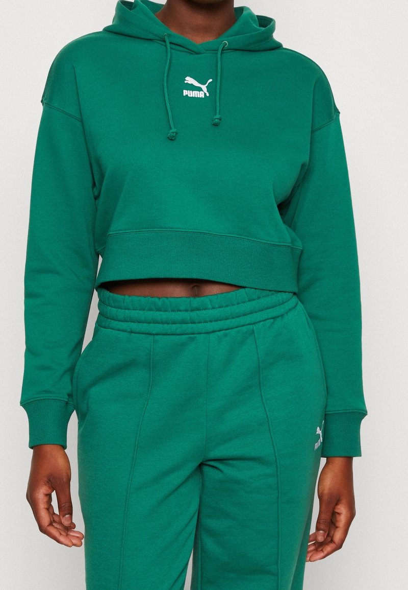 Sweat à capuche court vert avec cordon de serrage, arborant un logo Puma blanc, assorti à un pantalon de survêtement vert. Matière en mélange de coton doux.