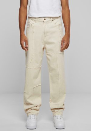 Untere Hälfte einer Person, die offwhite Patchwork-Hosen mit geradem Bein und weißen Sneakern trägt, stehend vor schlichtem grauem Hintergrund.