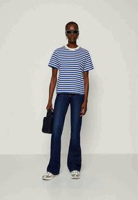 Stripete blå og hvit t-skjorte med rund hals, kombinert med mørke svingte jeans og sølvfargede sneakers; holder en liten svart veske.