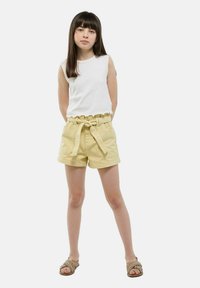 Haut sans manches blanc avec un ourlet volanté, associé à un short jaune avec une taille ceinturée et des poches avant. Le mannequin porte des sandales tressées.