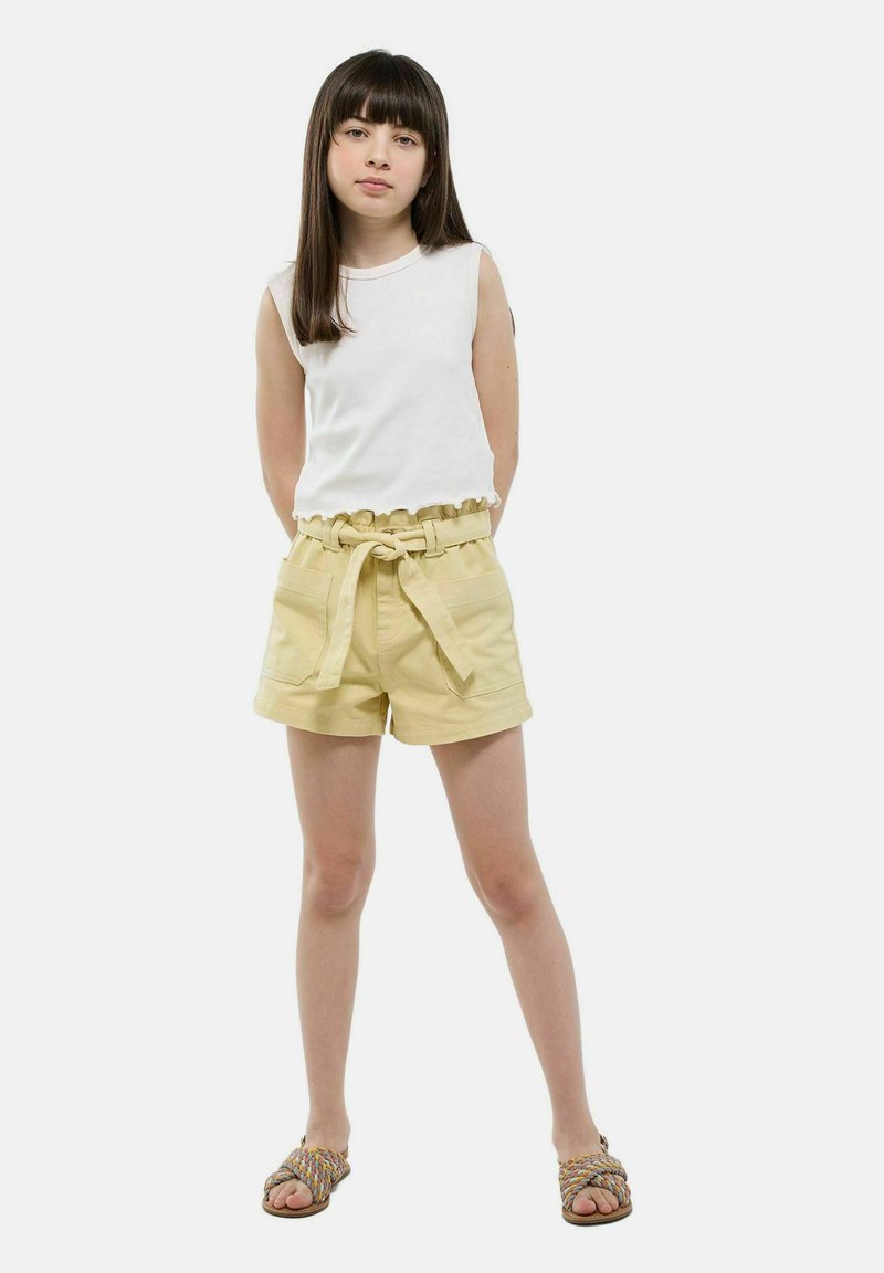 Haut sans manches blanc avec un ourlet volanté, associé à un short jaune avec une taille ceinturée et des poches avant. Le mannequin porte des sandales tressées.