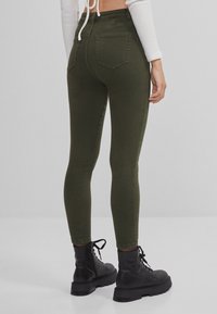 Femme portant un pantalon slim vert foncé, un haut à manches longues blanc court et des bottes de combat noires à lacets, debout devant un fond gris.
