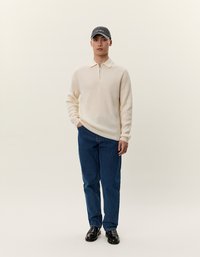Pullover a coste rigate color crema con colletto e zip, abbinato a jeans blu a gamba dritta e mocassini neri. La modella indossa un cappellino grigio.