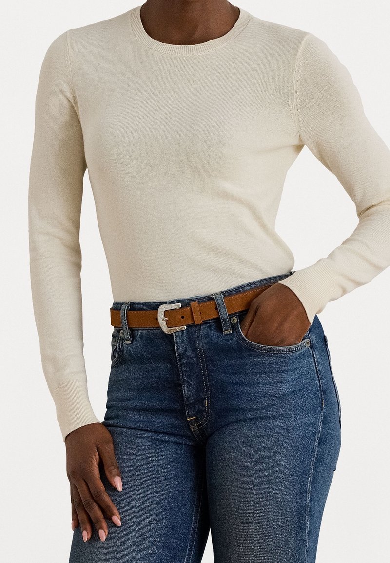 Personne à la peau foncée portant un pull à manches longues crème rentré dans un jean bleu taille haute avec une ceinture en cuir marron.
