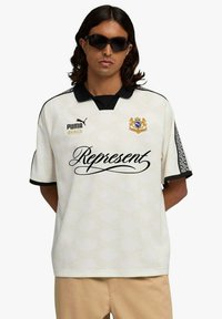 Camicia bianca a maniche corte con dettagli neri, caratterizzata da un colletto, logo sul petto e rifiniture con motivo intricato sulle maniche. Testo "Represent" sulla parte anteriore.