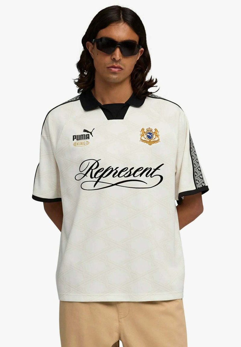 Camicia bianca a maniche corte con dettagli neri, caratterizzata da un colletto, logo sul petto e rifiniture con motivo intricato sulle maniche. Testo "Represent" sulla parte anteriore.
