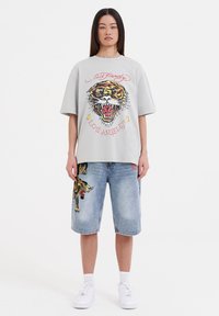 Grå oversized T-shirt med et tiger-motiv og rød tekst, parret med denimshorts med tigerbroderi på lommen. Hvide sneakers.