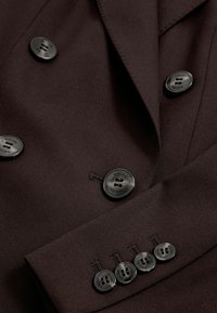 Blazer marron double boutonnage avec une texture de tissu lisse, comportant six boutons noirs avec le nom de la marque gravé et des revers ajustés.
