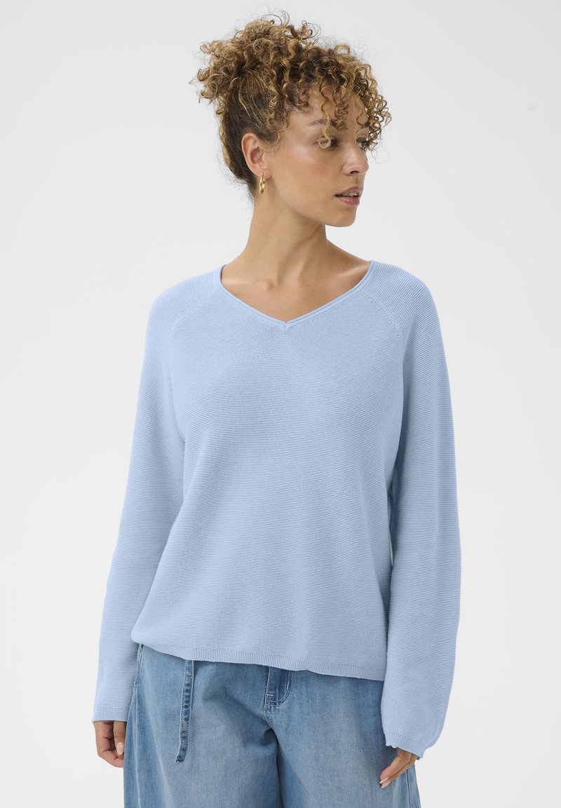 Femme aux cheveux bouclés portant un pull en V bleu clair et un pantalon en denim ample, se tenant devant un fond blanc uni, regardant vers la droite.