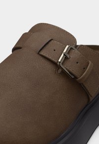 Καφέ σουέντ παπούτσι τύπου slip-on με μαύρη σόλα και λουράκι με γυαλιστερή μεταλλική πόρπη στο πάνω μέρος.