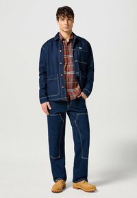Giacca di denim con cuciture bianche a contrasto, abbinata a una camicia di flanella a quadri in tonalità marroni e pantaloni in denim scuro, completata da stivali gialli.