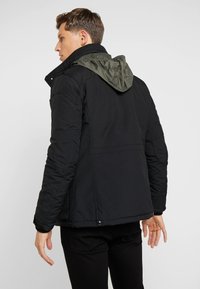 Black padded jacket with a green hood, featuring a sleek design, quilted texture, and snap button detail at the hem.