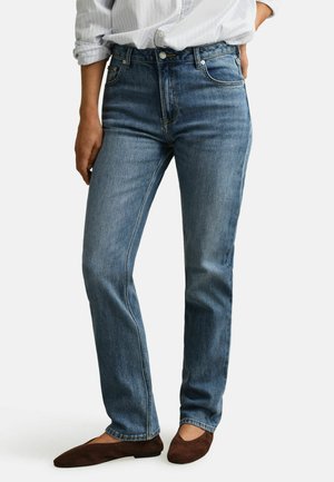 Personne portant un jean bleu taille haute à jambes droites, une chemise blanche rentrée et des chaussures plates marron, debout devant un fond blanc.