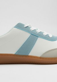 Zapatilla blanca con acentos en ante azul claro y una puntera beige. Presenta cordones redondos y una suela de goma texturizada con patrones diagonales.