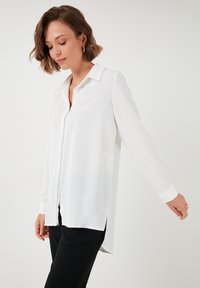 White long-sleeve shirt with a button-down collar, slightly longer back, and smooth texture, paired with black pants.