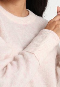 Frau, die einen weichen, hellrosa, flauschigen Pullover mit langen, gerippten Bündchen trägt und mit der rechten Hand ihren Kragen in der Nähe des Halses berührt.