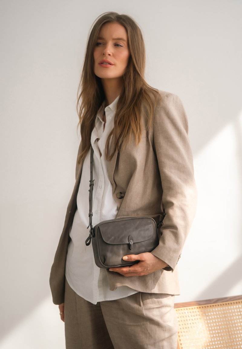 Borsa a tracolla in pelle grigia con chiusura a patta, design sottile, tracolla regolabile e dettagli di cucitura discreti, tenuta da una persona in giacca.