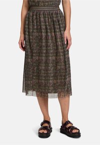 Gelaagde midi rok in donkergroen met een geometrisch patroon in gedempte roze en bruine tinten, voorzien van een stoffen tailleband en een doorschijnende onderrand. Zwarte sandalen.