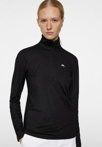 Musta vetoketjullinen pitkähiha toppi, jossa on teksturoitu geometrinen kuvio ja korkea kaulus; rinnassa pieni valkoinen logo.