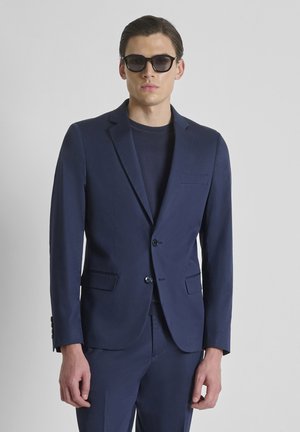 Man in een donkerblauw pak en trui met zwarte zonnebril, staand tegen een effen grijze achtergrond.