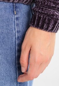 Manga de un suéter de terciopelo acanalado morado junto a una tela de denim azul claro, mostrando una textura suave y un diseño ajustado. Mano descansando contra los jeans.