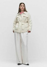 Krämfärgad oversized jacka med två framfickor, åtsydd midja och knappar, kombinerad med vida vita byxor och svarta skor.