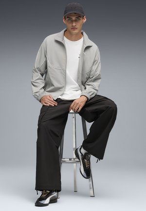 Ljusgrå zip-up jacka med elastiska muddar, vit t-shirt, lösa svarta byxor och svarta sneakers med färgglada detaljer, sittande på en pall.