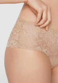 Braga de encaje en color nude con patrón floral, borde festoneado y textura suave y translúcida. Cintura elástica para mayor comodidad.