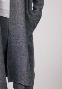 Personne portant un cardigan en laine gris clair avec une main dans la poche sur un pantalon assorti et un haut blanc, sur un fond neutre.