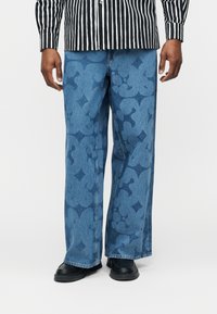 Άνδρας που φορά μπλε τζιν με μεγάλα λουλουδένια σχέδια και μαύρα παπούτσια, σε συνδυασμό με πουκάμισο με ασπρόμαυρες κάθετες ρίγες.