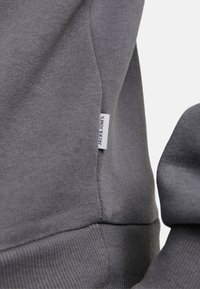 Sweatshirt gris avec des poignets et un ourlet côtelés, comportant une étiquette tissée sur le côté. Texture de tissu lisse avec un design ajusté.