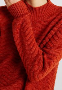 Pletený svetr v bohaté červené barvě s žebrovaným vysokým límcem a texturovanými vlnovými vzory na rukávech a těle.