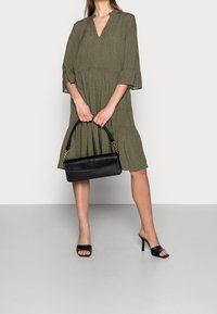 Žena v olivově zelených šatech s puntíky, držící černou kabelku a obutá v černých lodičkách na vysokém podpatku na neutrálním pozadí.