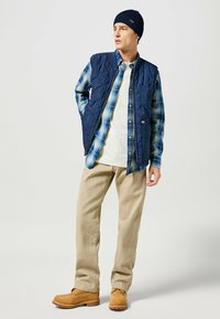 Navy quilted vest over a blue plaid shirt, white T-shirt, beige pants, and tan boots. The outfit features layering and textured materials.