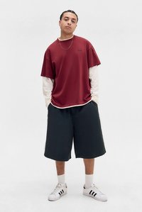 Jaunas žmogus stovi su kišenėse įkištomis rankomis, dėvi sluoksniuotą ilgomis rankovėmis baltą marškinį ir bordo spalvos trumpas rankoves, juodus šortus ir baltus sportinius batelius.