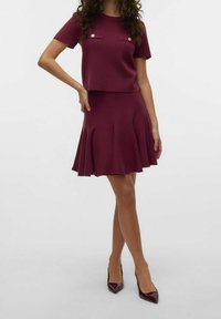 Top de punto burdeos con mangas cortas y dos bolsillos en el pecho, combinado con una falda acampanada con pliegues. Tacones brillantes de punta. 