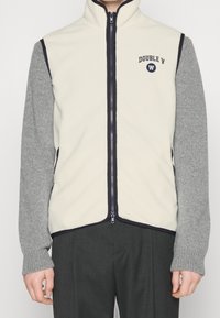 Chaleco de forro polar con cuello alto, parte delantera color crema, mangas grises, cierre de cremallera y detalles en azul marino. Presenta un logo bordado en el pecho.