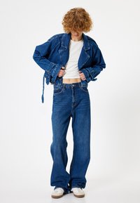 Chaqueta de mezclilla con solapas anchas y mangas largas, llevada sobre una blusa blanca y combinada con unos vaqueros azules de corte holgado. Zapatillas blancas abajo.