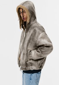 Veste à capuche grise, avec une finition texturée et une doublure en fausse fourrure beige. Comprend une fermeture éclair et des poches latérales. Design mi-crop.