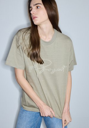 Frau mit langen braunen Haaren in hellgrünem, übergroßem T-Shirt mit perlenartiger Schrift, hält den Saum, trägt hellblaue Jeans, vor grauem Hintergrund.