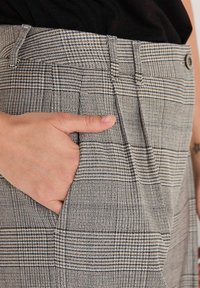Hände in die Seitentasche einer beigen und schwarzen karierten hochgeschnittenen Hose mit Knopf und Falten gesteckt.