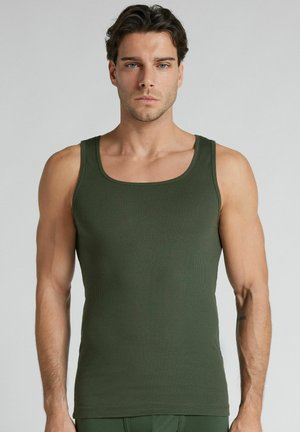 Mężczyzna z krótkimi ciemnymi włosami, ubrany w dopasowaną oliwkowozieloną prążkowaną koszulkę na ramiączkach, stojący na jednolitym jasnoszarym tle.