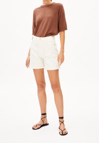 Henkilö, jolla on päällään ruskea ribbineulottu lyhythihainen paita, valkoiset käännetyt shortsit ja mustat remmillä varustetut tasapohjaiset sandaalit valkoisella taustalla.