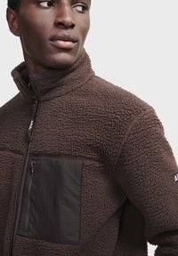 Brun fleecejakke med struktureret overflade, der har en sort stofbrystlomme og lynlåslukning, høj krave og minimalistisk design.