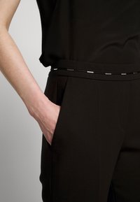 Černé kalhoty s hladkým povrchem a boční kapsou. Na pase je tenký dekorativní pásek s kovovými akcenty.